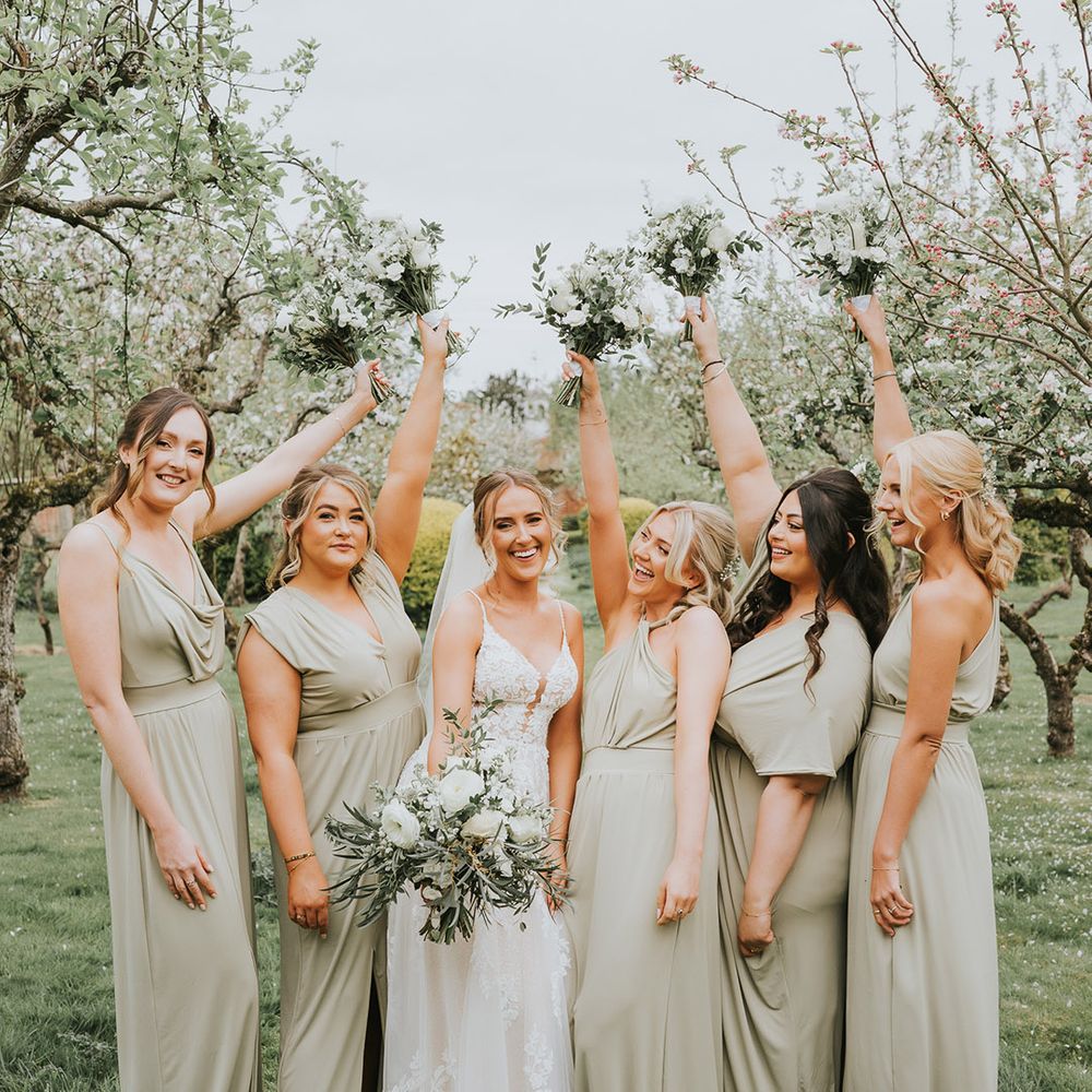 Bridesmaids in sage green dresses with the bride lifting up their white flower bouquet 