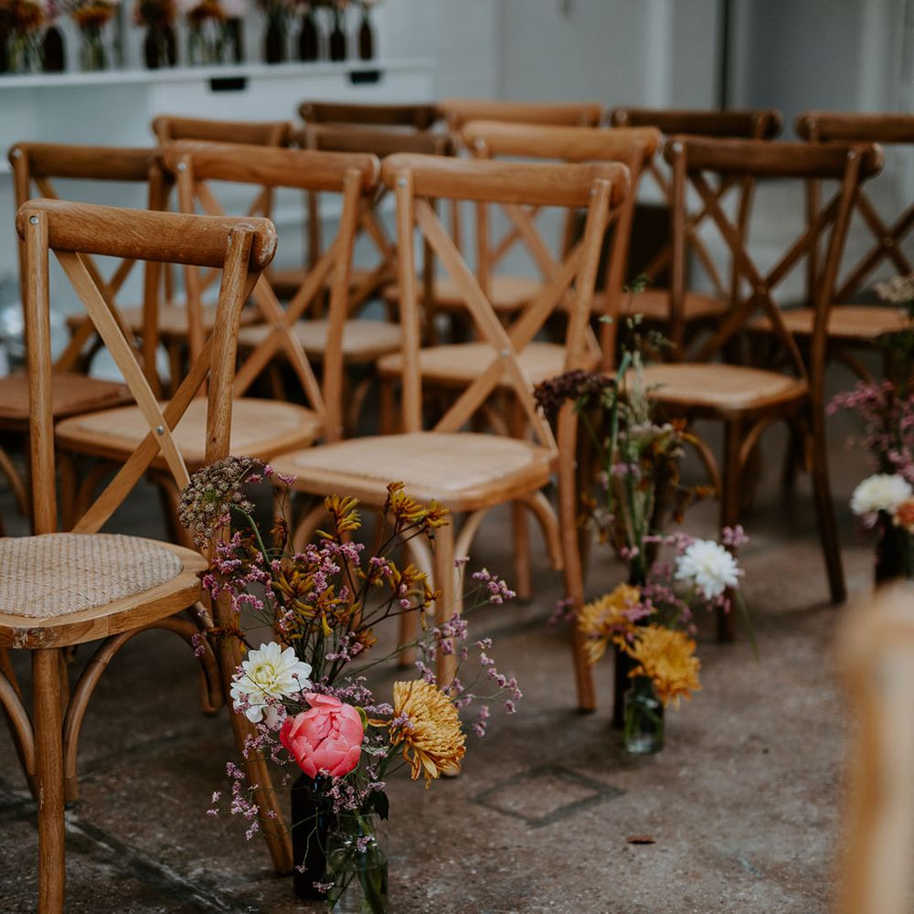 Small floral arrangements in bright colours beside wooden chairs lining the aisle