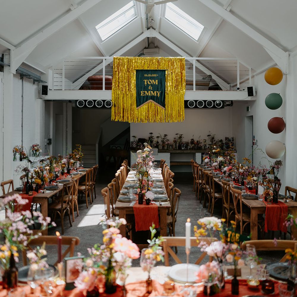 Industrial styled wedding decor complete with gold streamers and wooden banquet tables with orange fabric table runner and small floral arrangements
