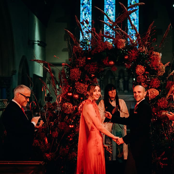 Bride in satin red wedding dress with cape and groom in three piece suit with red tie at winter wedding holding hands at alter