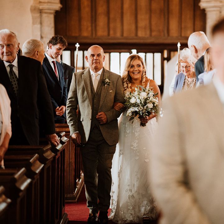 Father of the bride in a dark grey suit walks the bride in a lace wedding dress down the aisle to the groom waiting 
