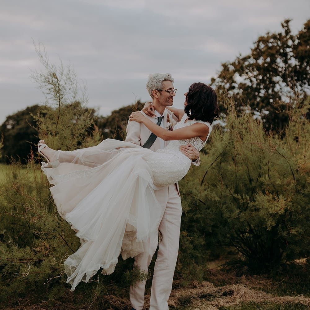 Groom in light pink suit carries bride wearing lace and tulle fishtail dress in front of greenery