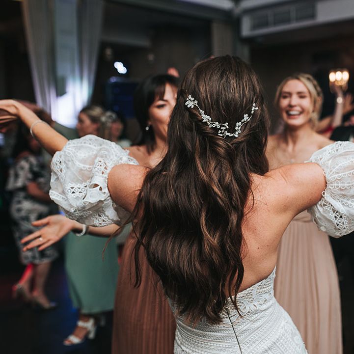 Bride wears her hair in a half up half down style with white flower accessory in a strapless lace gown with detachable sleeves dancing on the dance floor 
