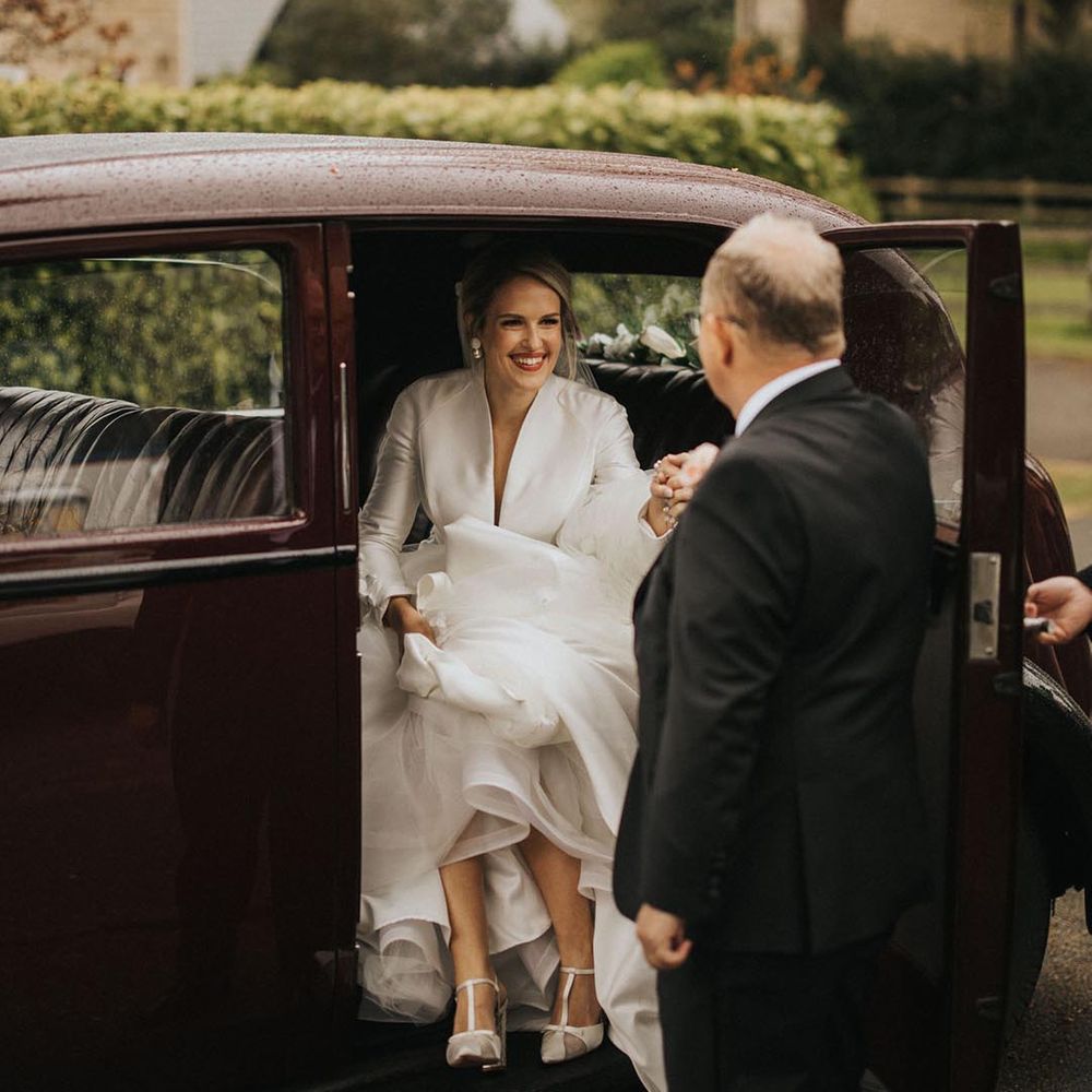 Father of the bride in black tie helps the bride out of the traditional vintage black wedding car in a conservative Jesus Peiro wedding dress