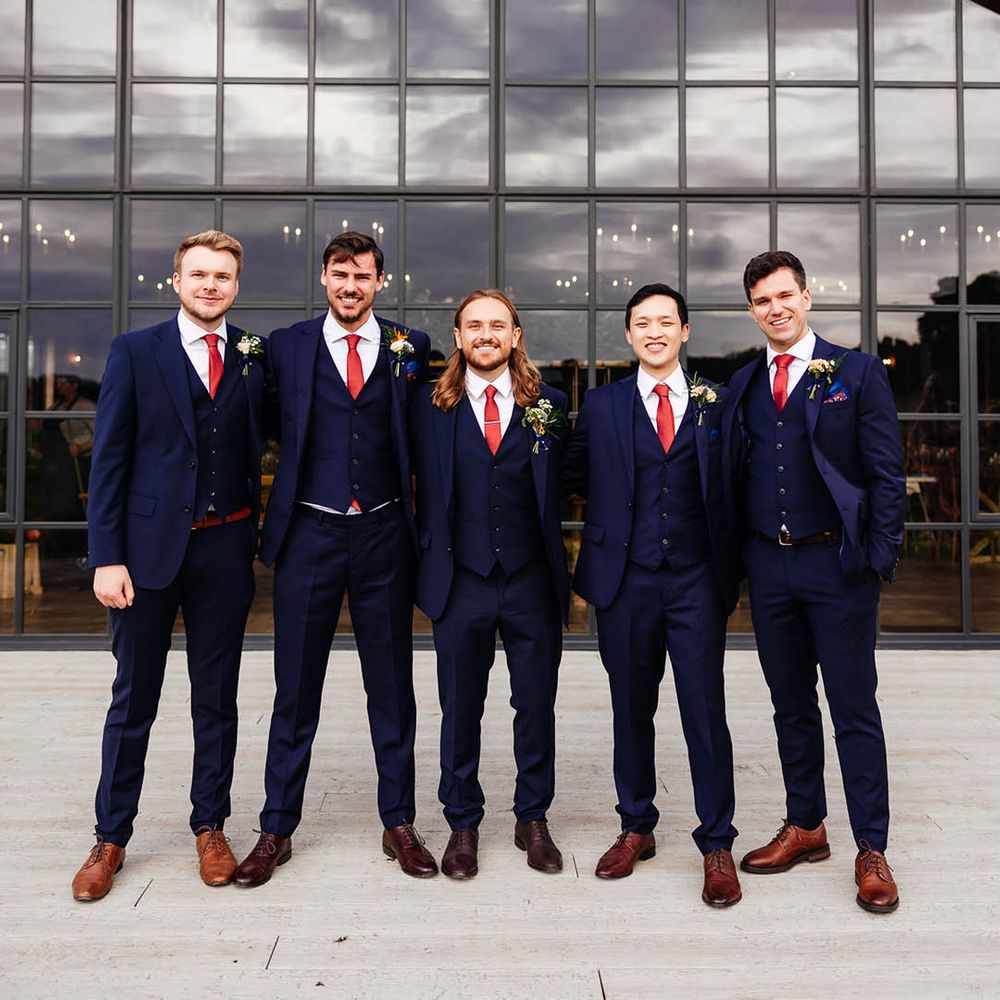 Groomsmen in matching blue three piece suits with red ties, brown shoes, and colourful buttonholes