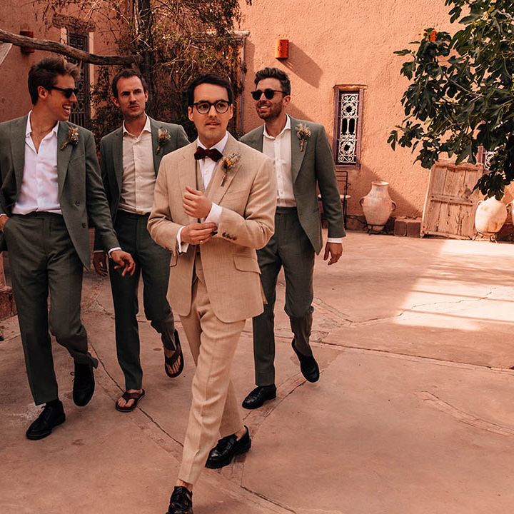 Groomsmen in dark grey suits with no ties walking with the groom in cream suit for summer destination wedding 