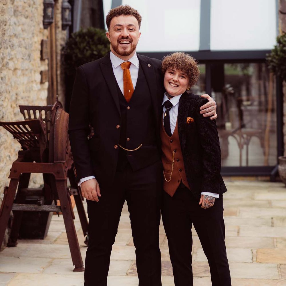 Bride in dark velvet bridal jacket with rust waistcoat and pocket square, black suit trousers, platform Dr Martens and gold pocket watch standing with wedding guest in dark suit with rust orange tie at Kingscote Barn Tetbury