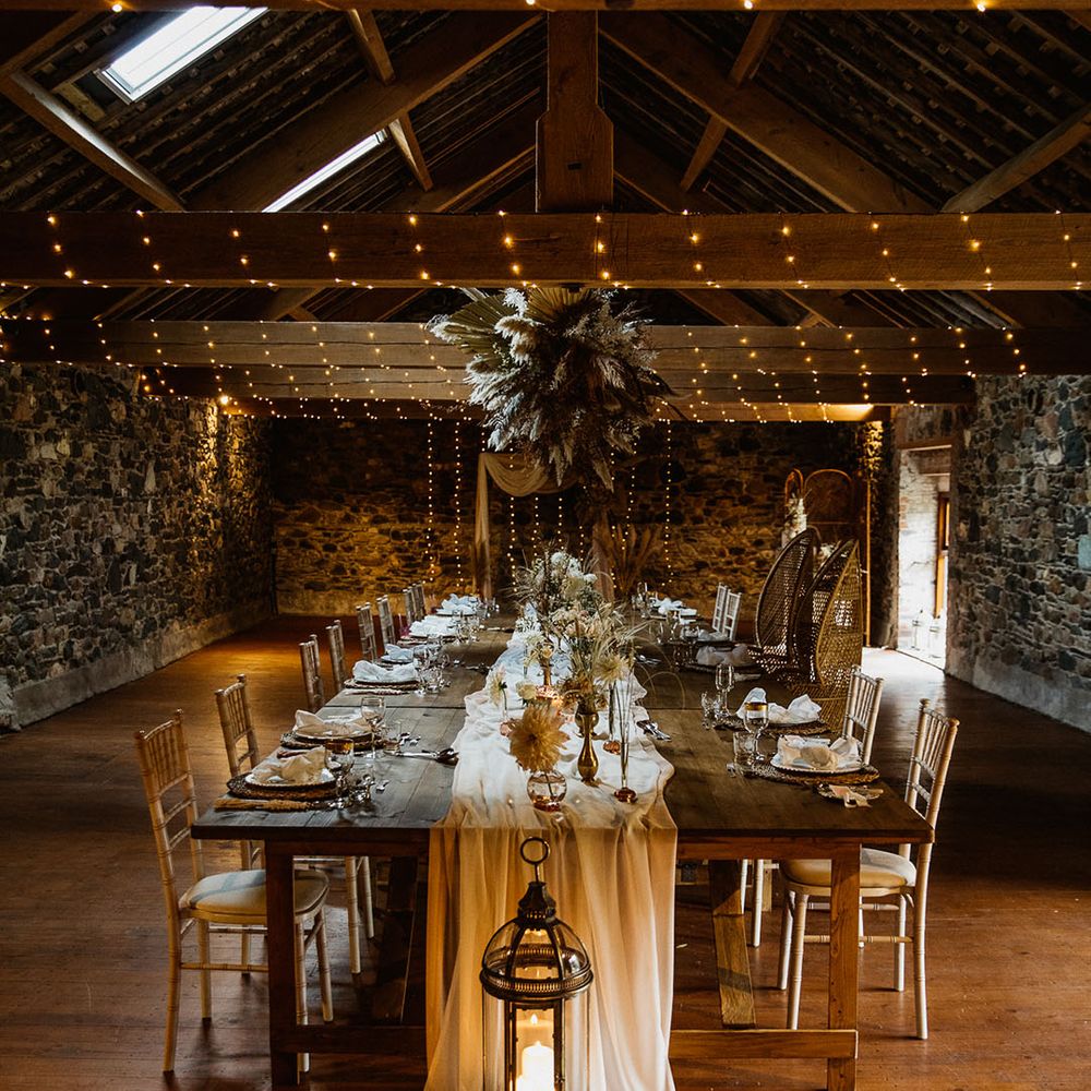 Wooden tables covered in white table runner and warm candlelight with pampas grass installation and dried wedding flowers 