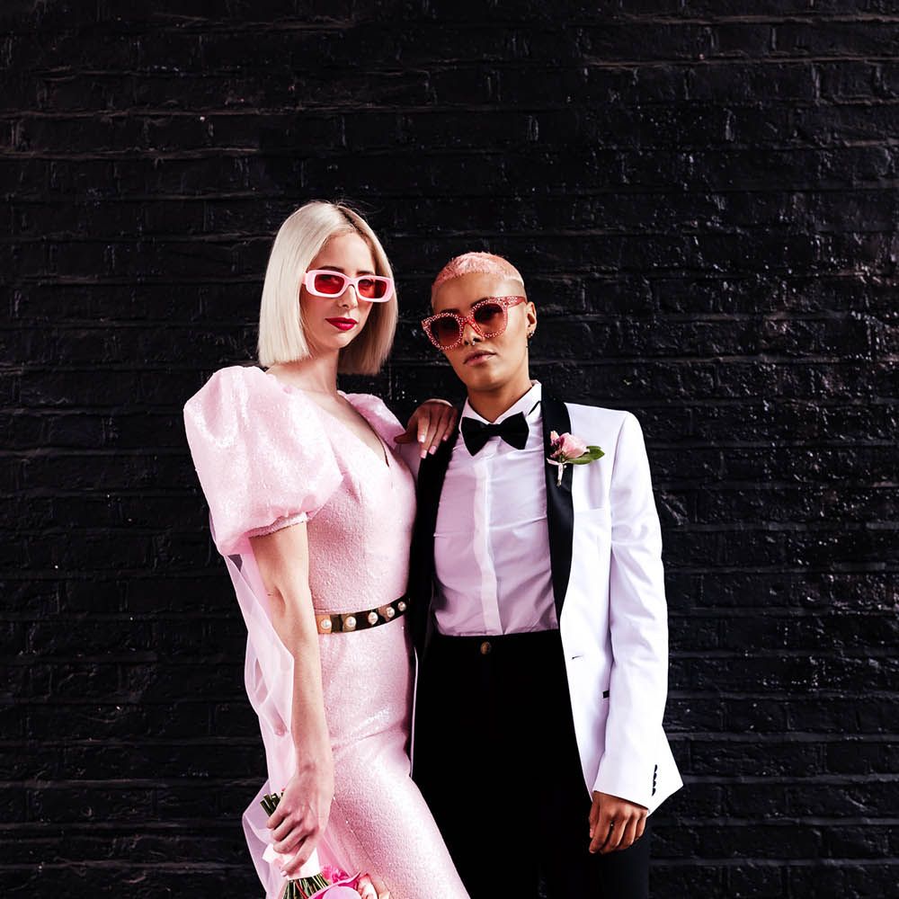 Alternative wedding fashion with bride in a pink jumpsuit and sunglasses and bride in a white tuxedo jacket with black lapel and bow tie 