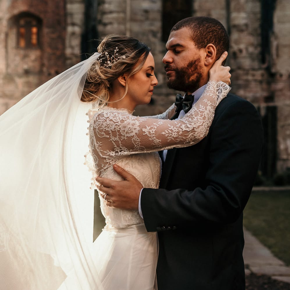 Bride and groom portrait with bride in a lace long sleeve wedding dress holding her grooms head as her appliqué veil blows in the wind 