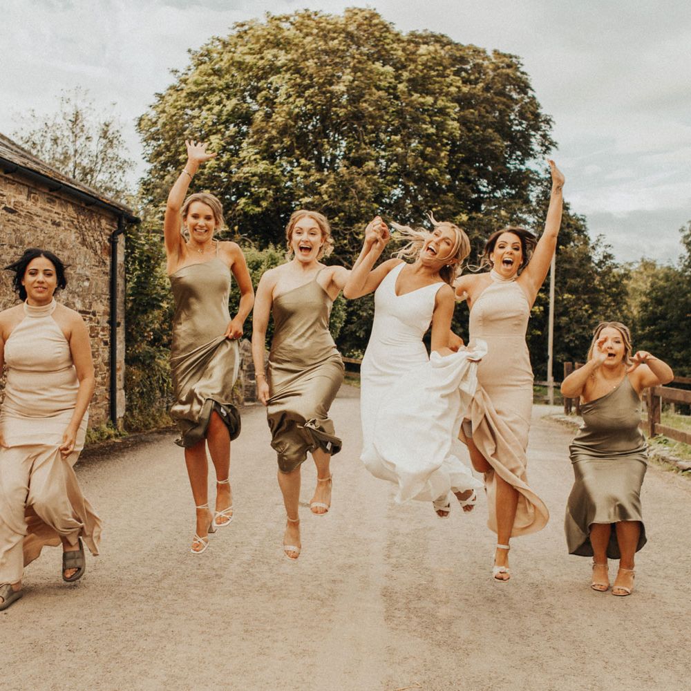 Bride in white Made With Love wedding dress and bridesmaids in sage green and beige bridesmaids dresses jump in the air on country lane at Anran Devon