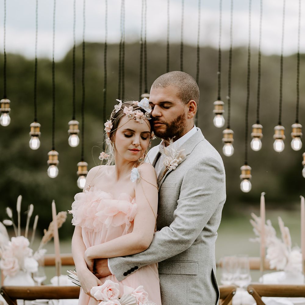 Groom in a grey suit embracing his bride in a blush pink wedding dress in front of a wedding reception table with Edison bulb light installation