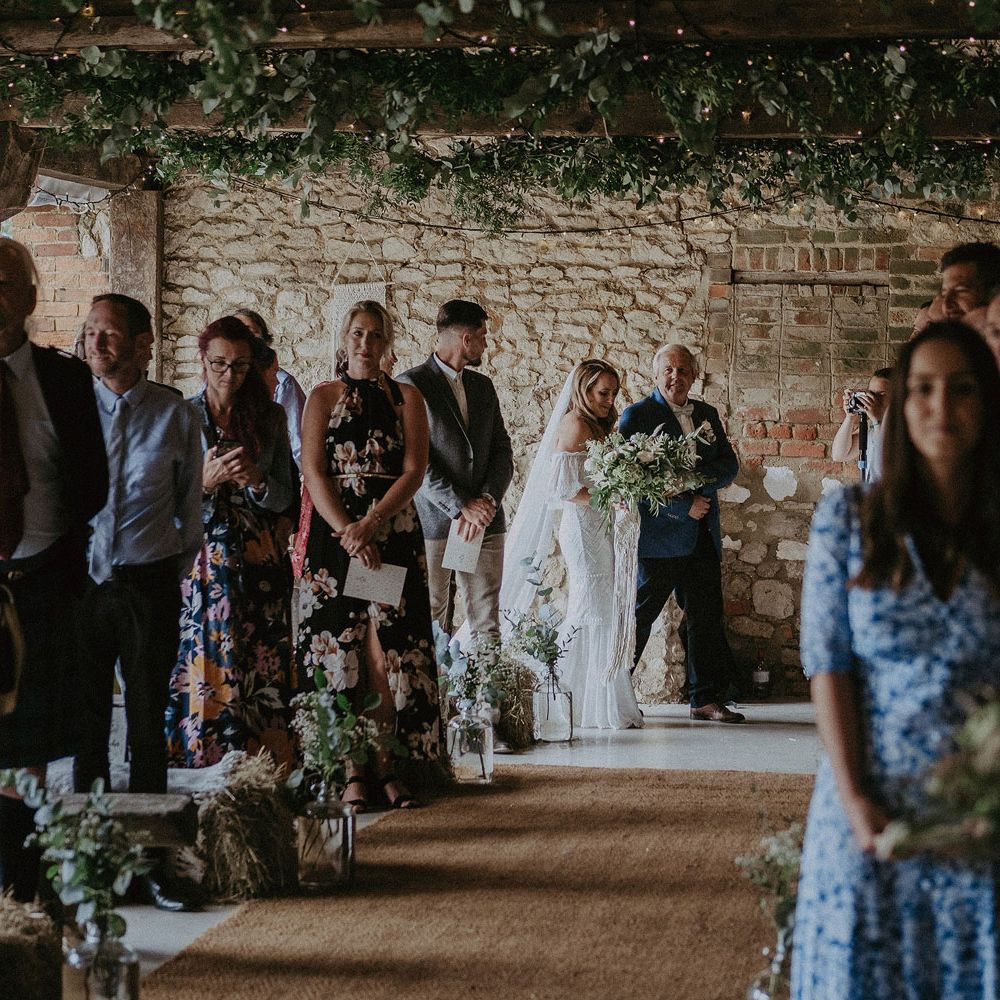 Bride in white Grace Loves Lace bardot wedding dress and veil holding white and green bridal bouquet walks into wedding ceremony with father in blue suit at Isle of Wight wedding with macrame wedding decor
