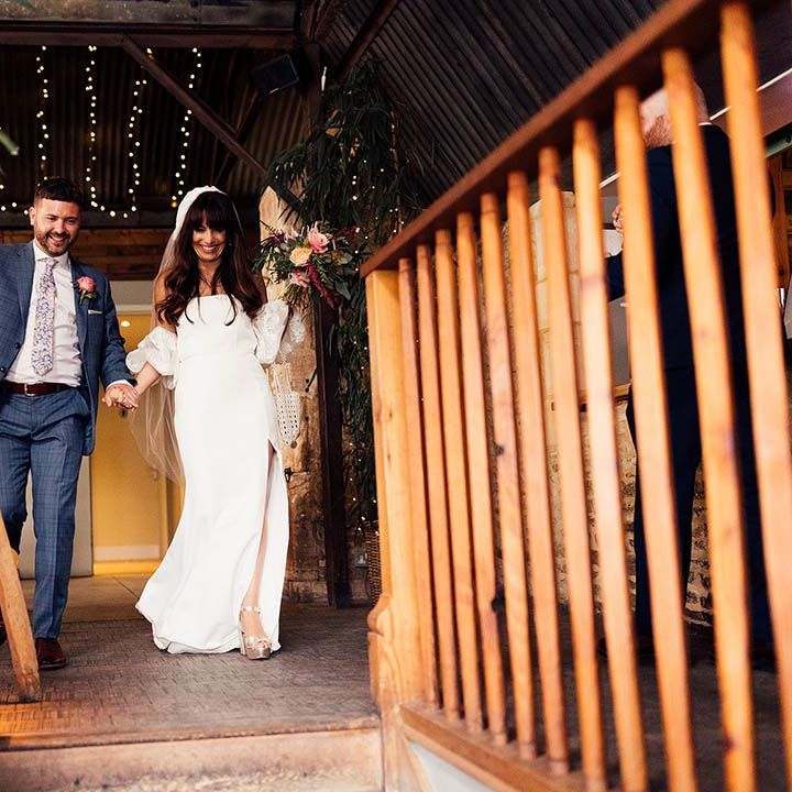 Bride in a wedding dress with front split showing off her gold Miu Miu platform shoes entering the wedding reception with her groom in a navy check suit 
