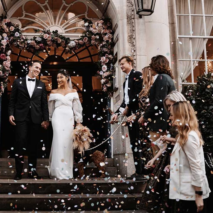 Wedding party throw confetti with joy and smile as bride and groom walk out of Chelsea Town Hall 