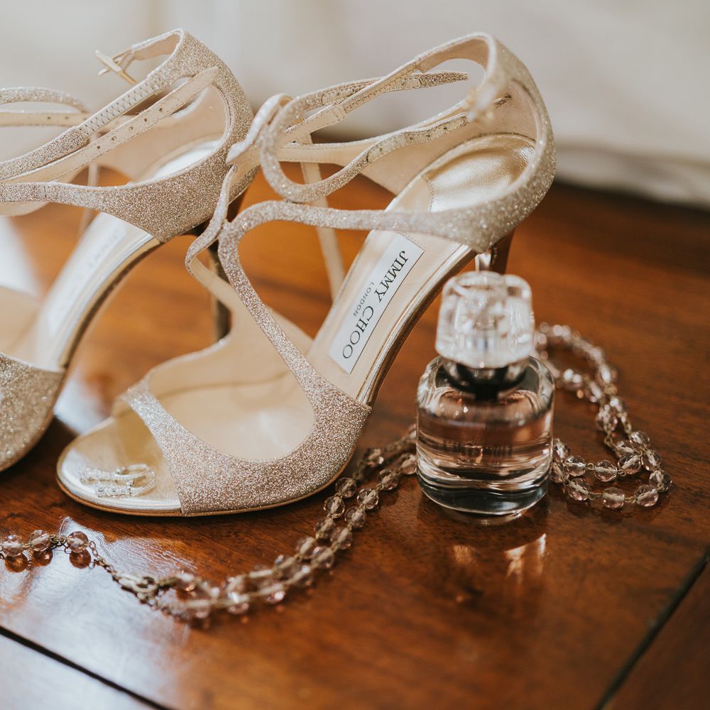 Gold sparkle strap Jimmy Choo wedding shoes with a bottle of perfume and rosary necklace on wooden table