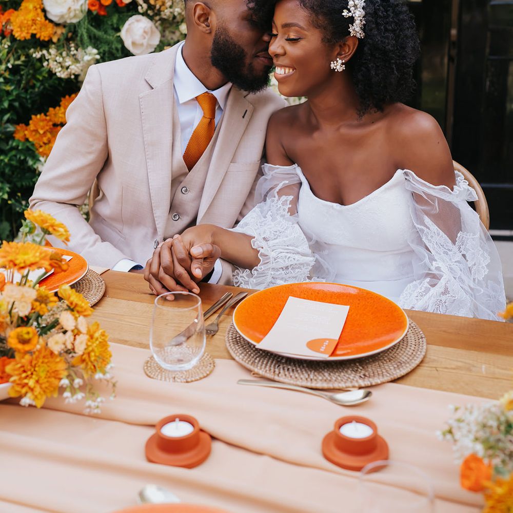 Groom in a beige suit and orange tie holding hands with his bride in a strapless wedding dress with lace sleeve at their orange outdoor wedding reception 