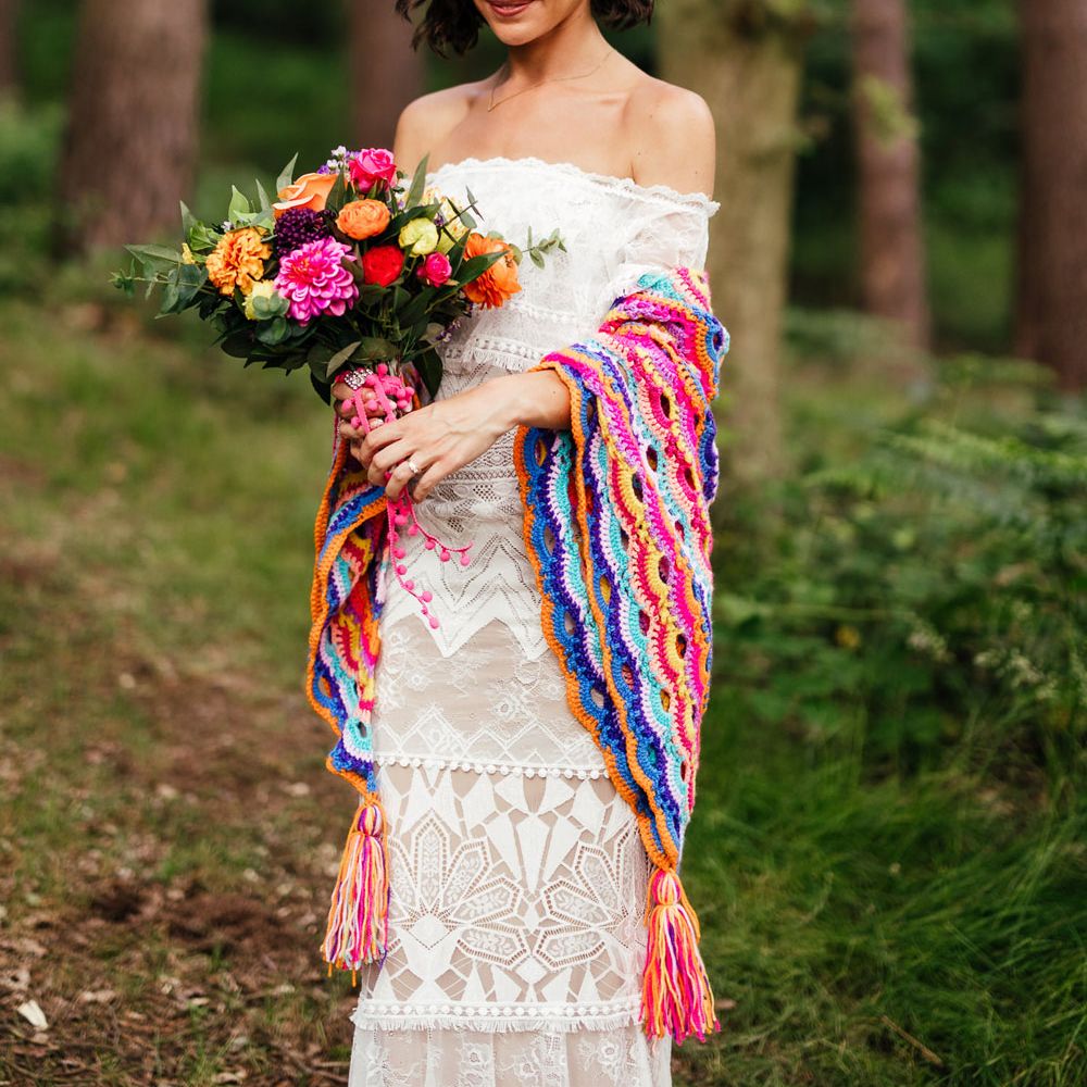 Smiling bride in off the shoulder Grace Loves Lace wedding dress and colourful flower crown holds colourful bridal bouquet and wears colourful crocheted blanket as she stands in woodland after outdoor wedding ceremony