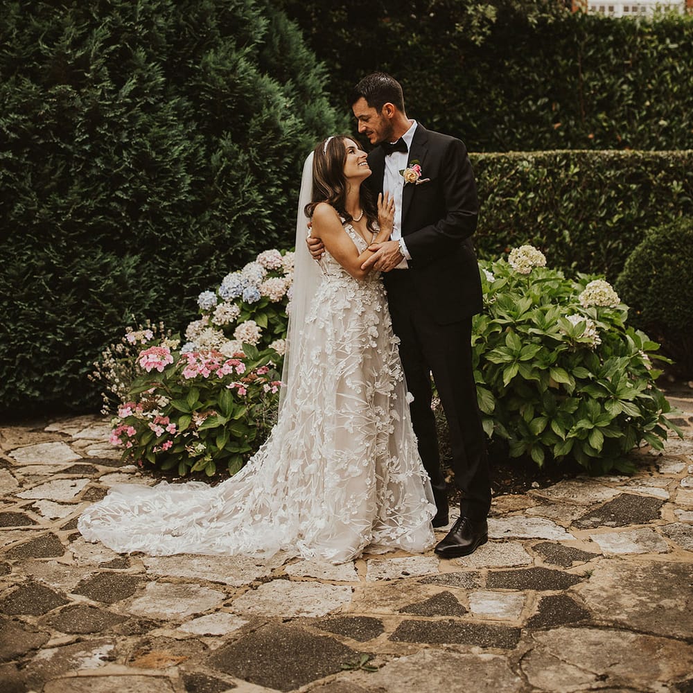 Bride leans into her groom as they stand outdoors in garden on their wedding day
