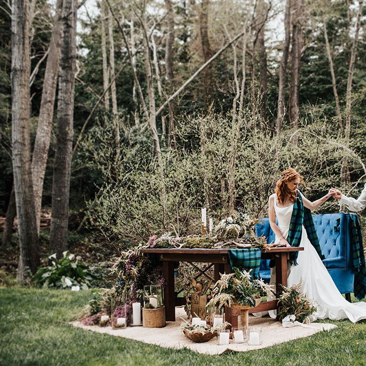 Groom in a blue and green wedding kilt holding hands with his bride in an A line wedding dress at their outdoor boho sweetheart table reception 