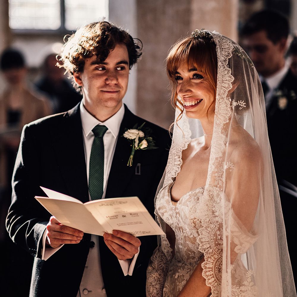 Groom gazes at bride as she beams during church ceremony in lace trim veil