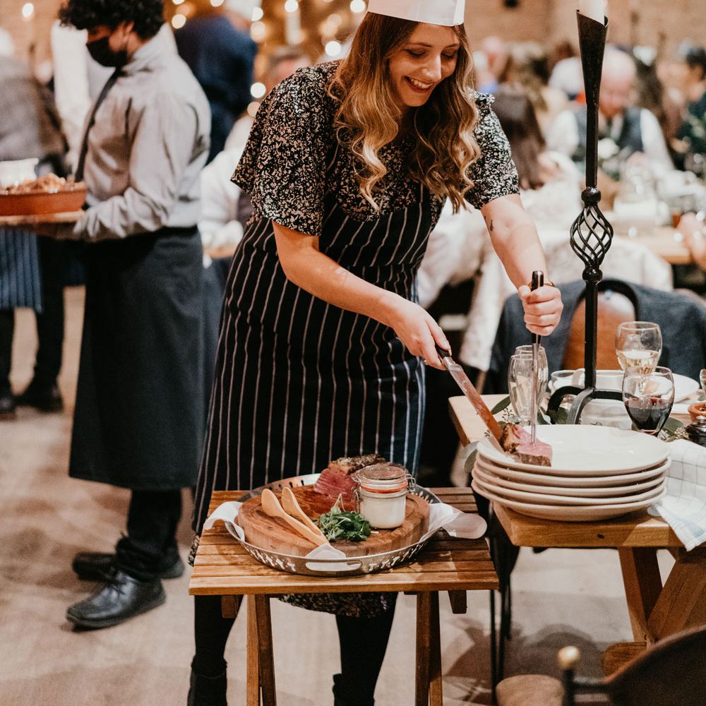Guests in striped apron and chefs hat serves meat to a plate during barn wedding breakfast at The Tythe Barn in Launton