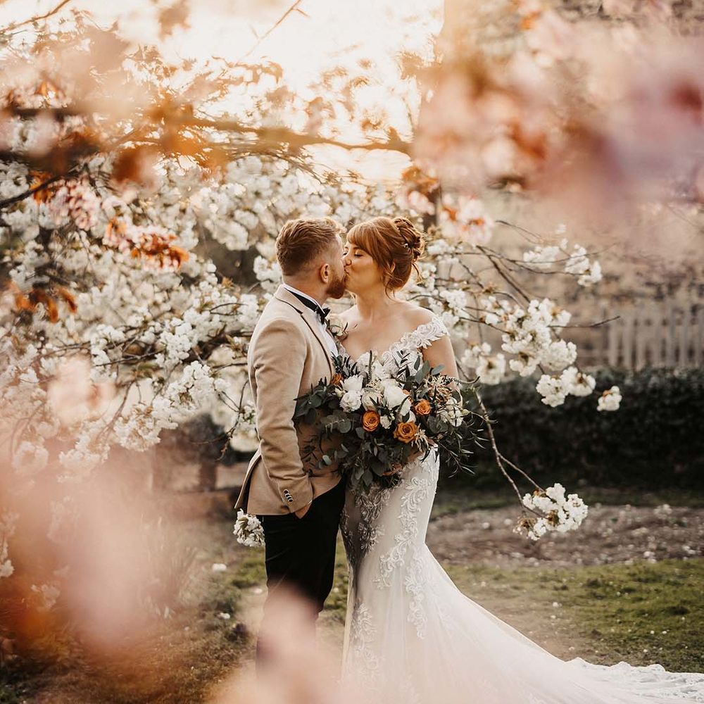 Bride and groom share a kiss under the blossom trees for spring wedding in rustic barn venue