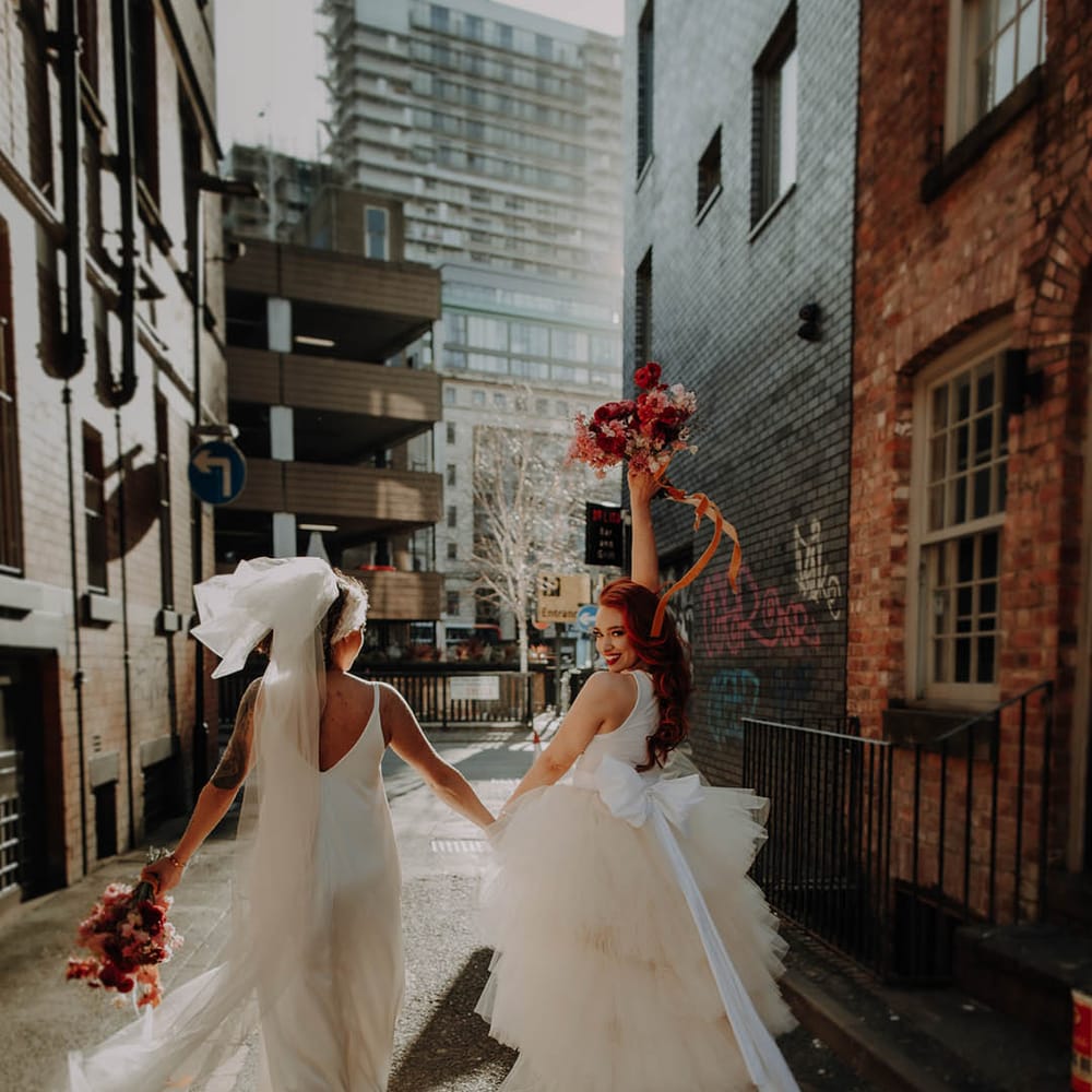Manchester elopement with two brides in a layererd tulle wedding dress holding a red bouquet in the air and a satin slip wedding dress long bow veil 