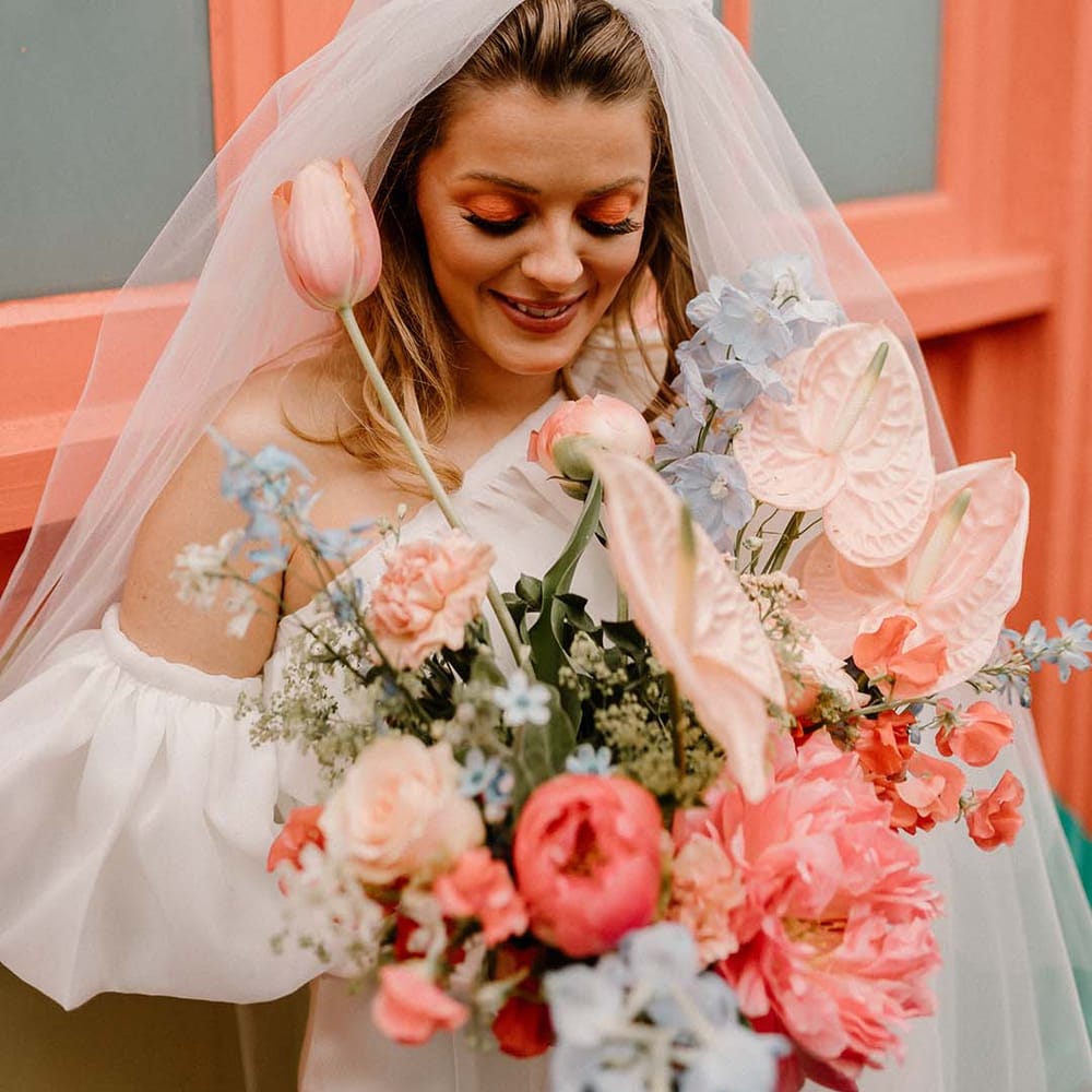 Bride with coral eye shadow wedding makeup and false eyelashes holding a pink and blue wedding bouquet with anthuriums, peonies and delphiniums 