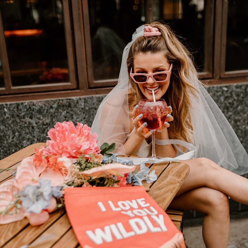 Bride in retro pink sunglasses and a short wedding dress drinking a cocktail with a pink suede fabric wedding banner on the table 