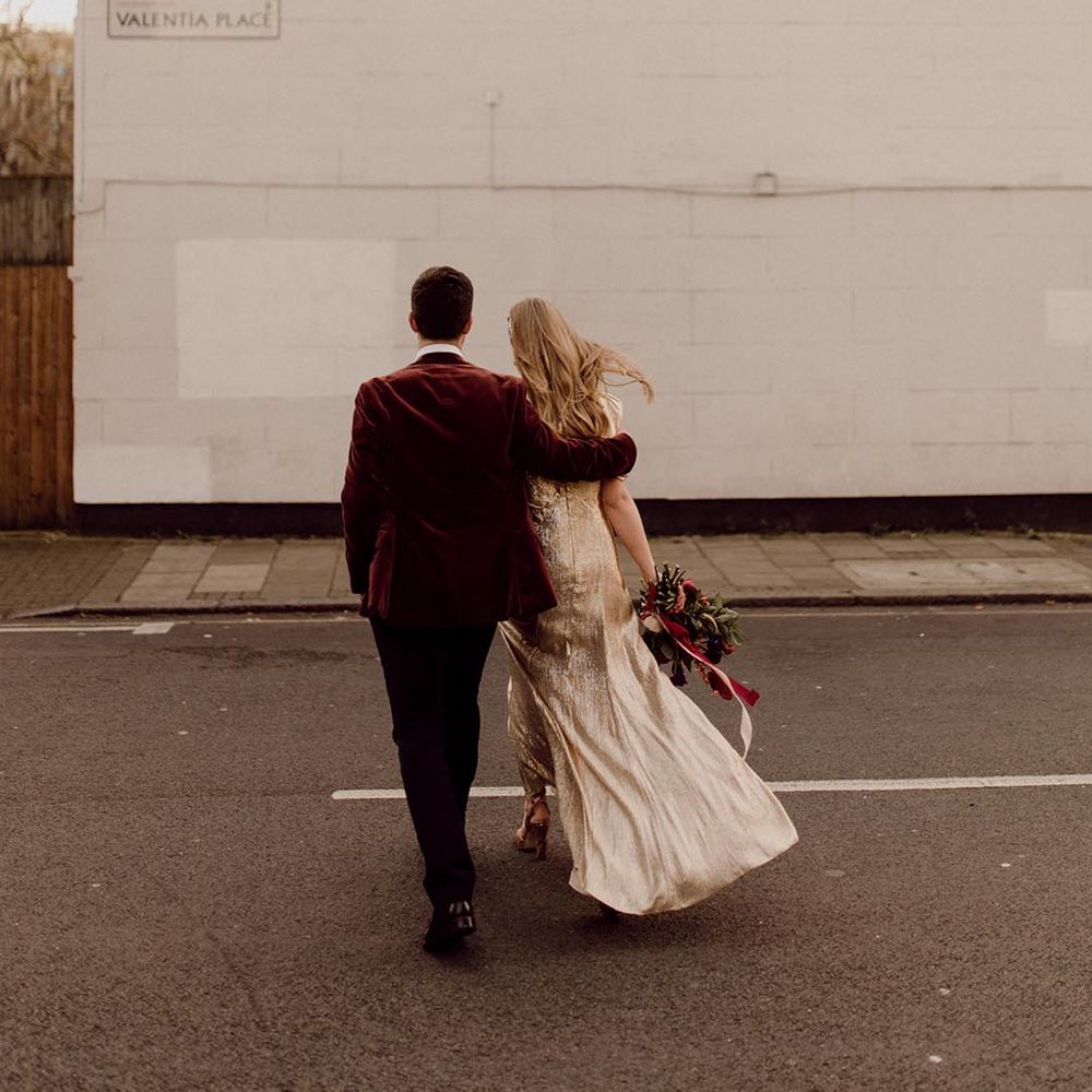Groom in velvet suit jacket holds the bride in sparkly gold wedding dress walk around their city wedding venue together 