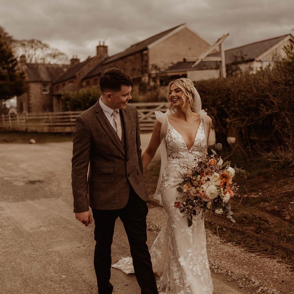Bride in v neck lace wedding dress with bow sleeves walking with the groom in brown suit holding orange and white bouquet