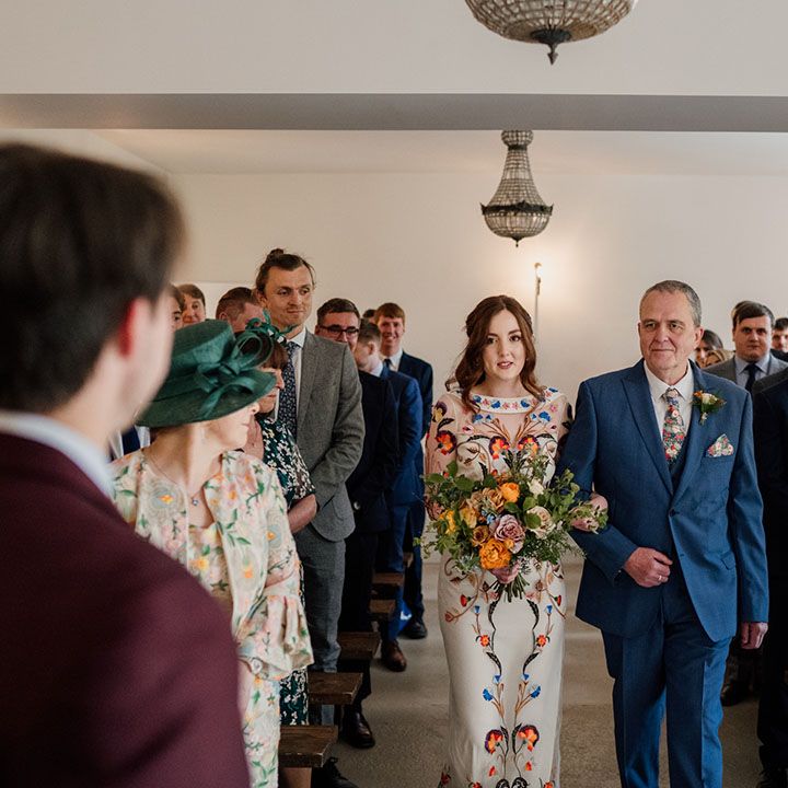 Father of the bride in dark blue three piece suit with bride in Temperley Bridal dress heads towards the groom in burgundy suit
