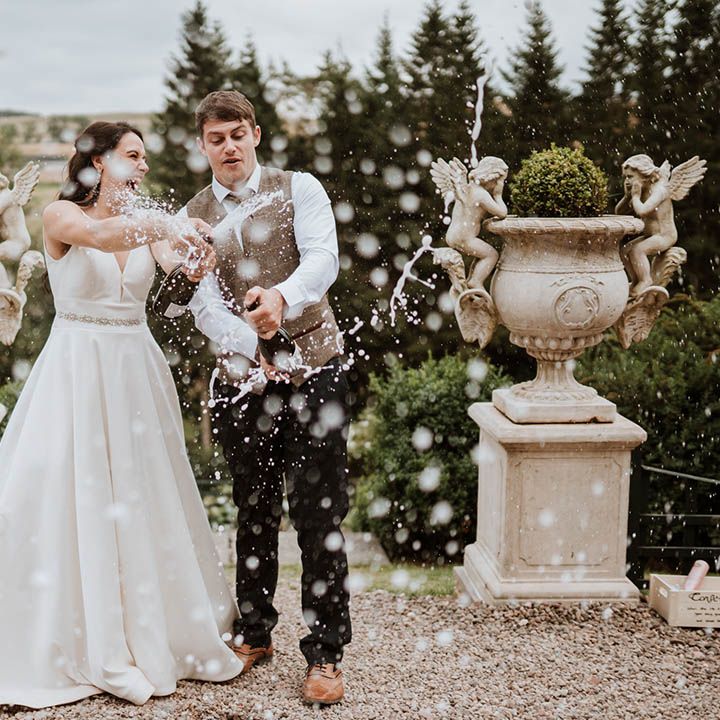 Bride a princess wedding dress and groom in a wool waistcoat popping champagne at their country house wedding 