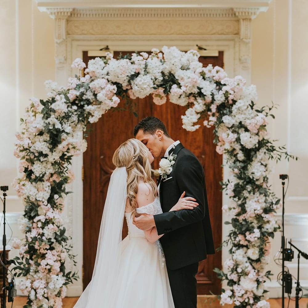 Bride and groom share their first kiss as a married couple in front of pink and white flower arch 