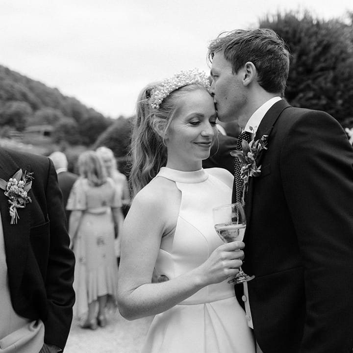 Groom in morning suit kisses the bride on the forehead as she sips from a glass 