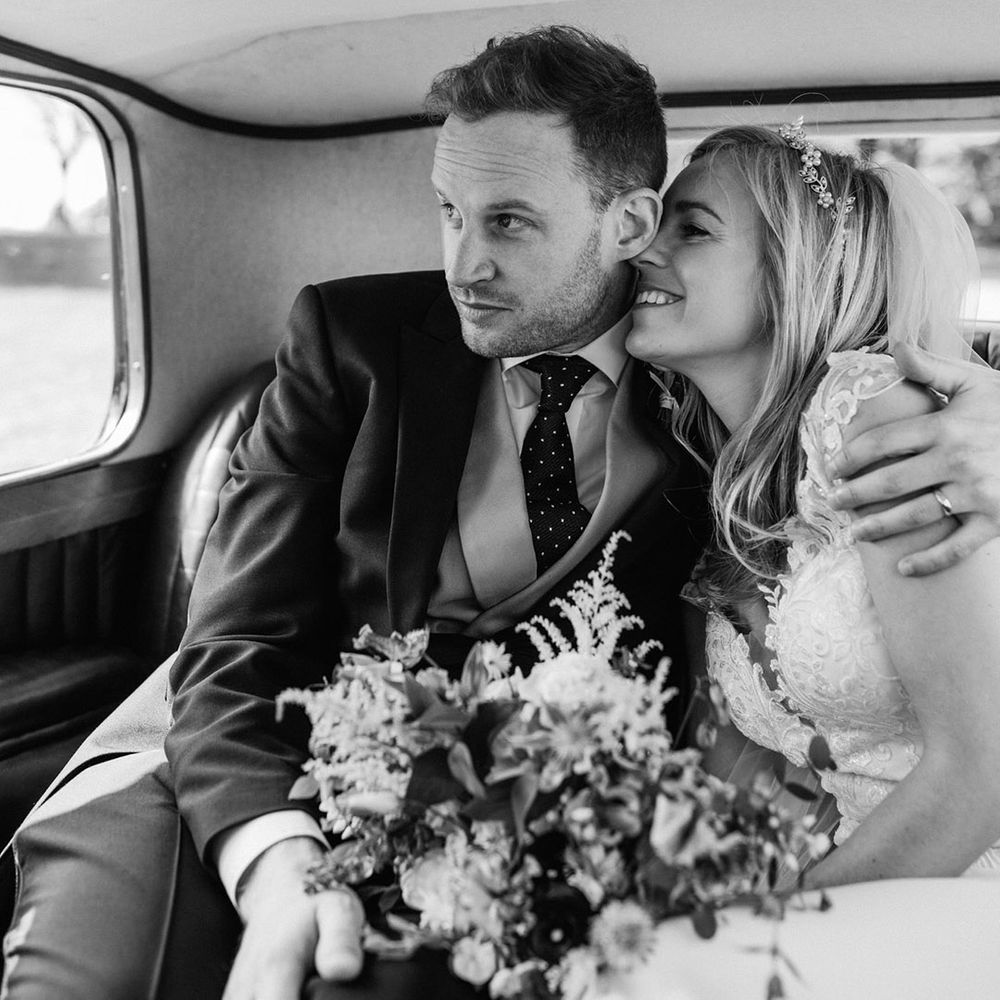Bride in lace wedding dress wearing pearl and gold headpiece with groom in morning suit seated in vintage wedding car 