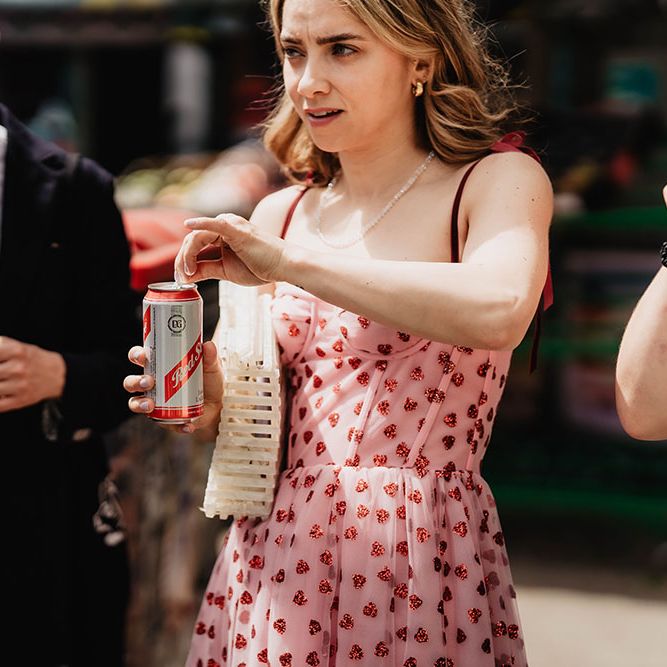 Wedding guest in pink dress complete with glittery spots opens can of beer outdoors in the sunshine 