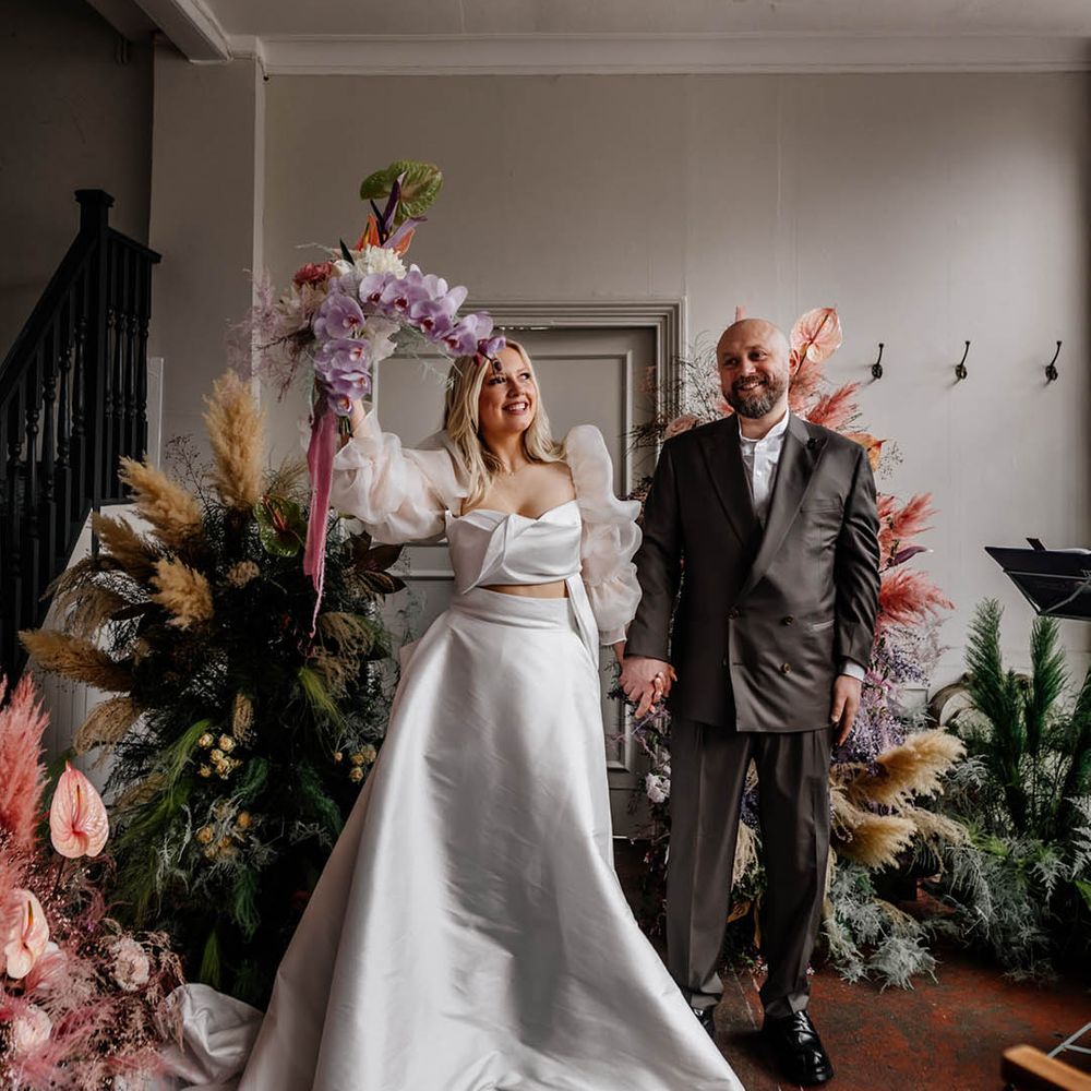 Groom in a double breasted suit with his bride in a JESUS PEIRO wedding dress holding her exotic flower bouquet in the air 