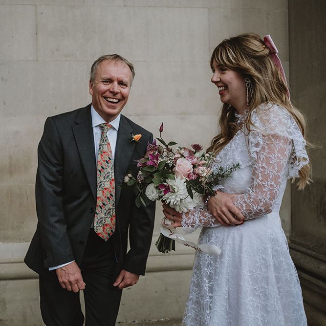 Bride with pink fabric bow in her long blonde hair carries sustainable floral bouquet whilst wearing lace vintage wedding dress with long sleeves