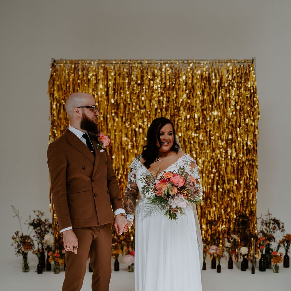 Groom wears 70s style retro brown suit and stands beside his bride in lace wedding dress holding brightly coloured floral bouquet 