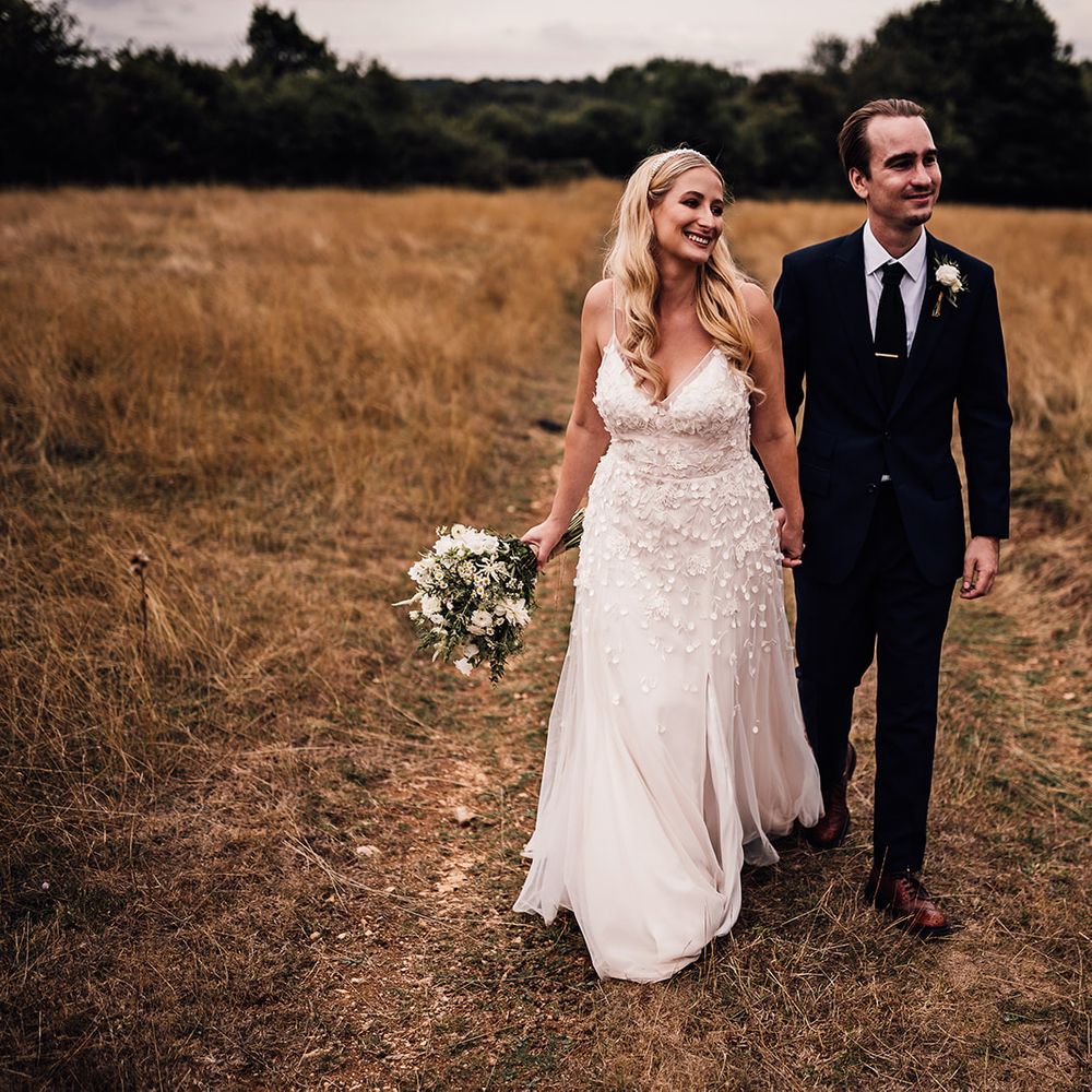 Bride in a flower appliqué Alexandra Grecco Lana wedding dress walking in the fields with the groom in a navy blue suit 
