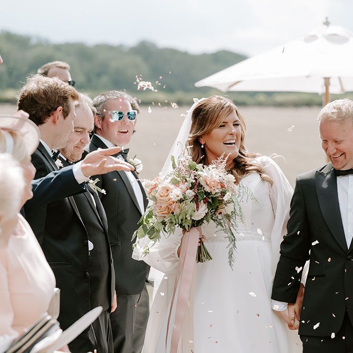 Bride in romantic wedding dress walks through white petal confetti with her groom in black tie