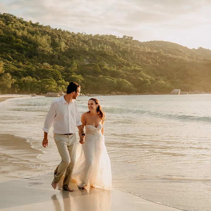 Bride in off the shoulder lace wedding dress with sweetheart neckline and lace floral details holding hands and laughing with  groom in crisp white shirt and light coloured chinos walking along the beach
