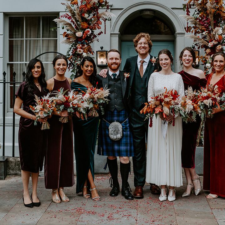 Bride in long sleeve lace vintage wedding dress, groom in dark suit with orange tie and autumnal boutonniere, best man in dark blue kilt and bridesmaids in mismatched velvet bridesmaid dresses standing underneath large floral arch with roses, peonies, foliage and pampas grass