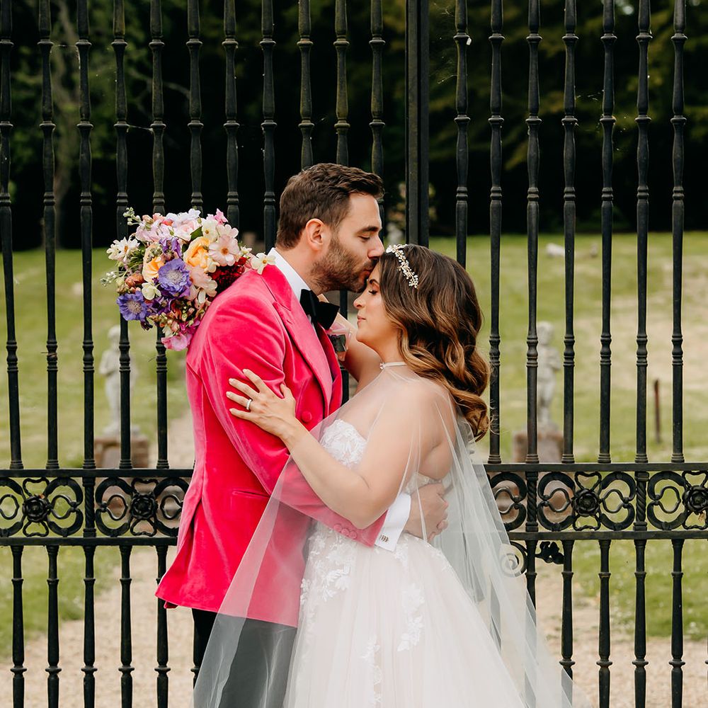 The groom in a bright vibrant wedding outfit kisses the bride on the forehead as she holds a colourful wedding bouquet 