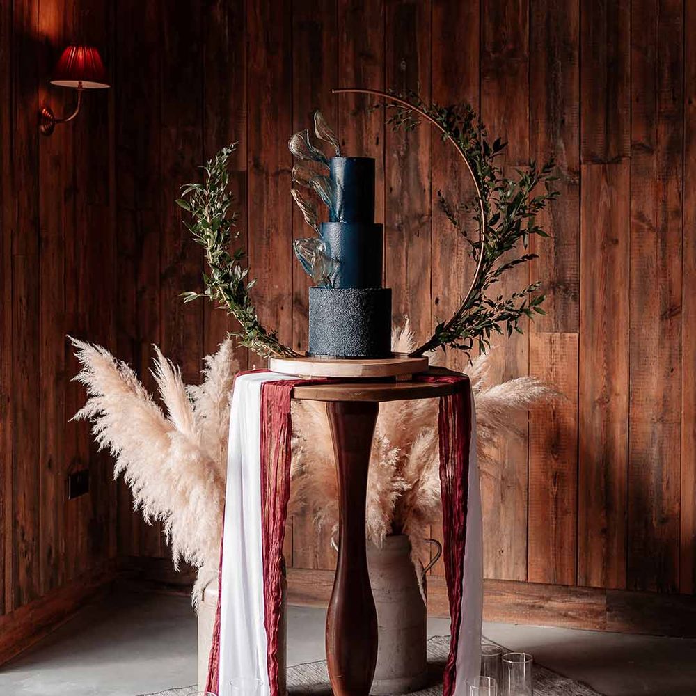 three tier wedding cake in a rich blue buttercream with a velvet effect base tier and gold rice paper sails, finished with a semi circle Bramble Sky arch stand and decorated with lots of dark green ruscus 