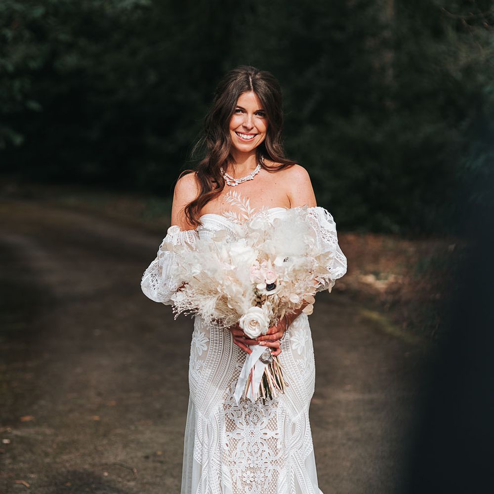 Bride in boho fitted wedding dress with a train and detachable sleeves holding an all white fired flower bouquet tied with white ribbon for boho luxe wedding 