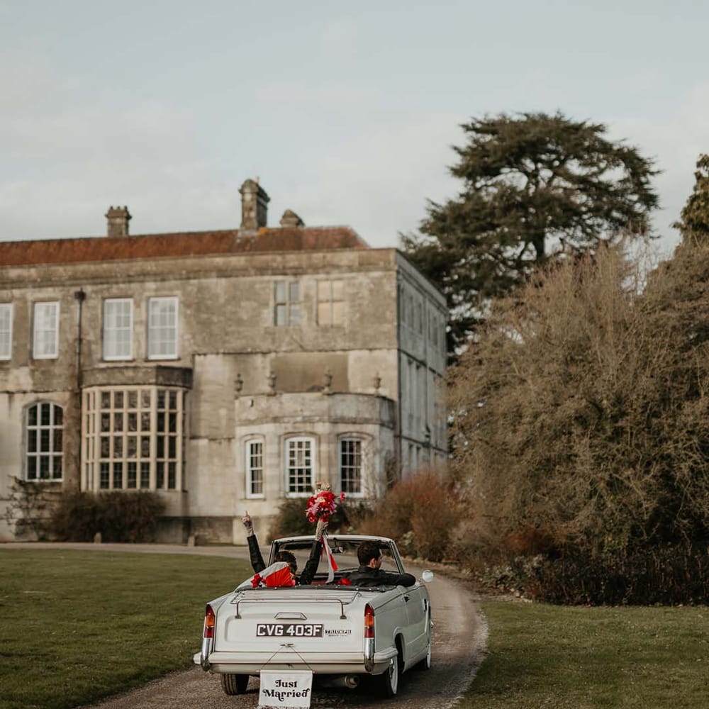 Couple driving to Elmore Court wedding venue in off white classic convertible wedding car with white fabric “just married”  banner decoration
