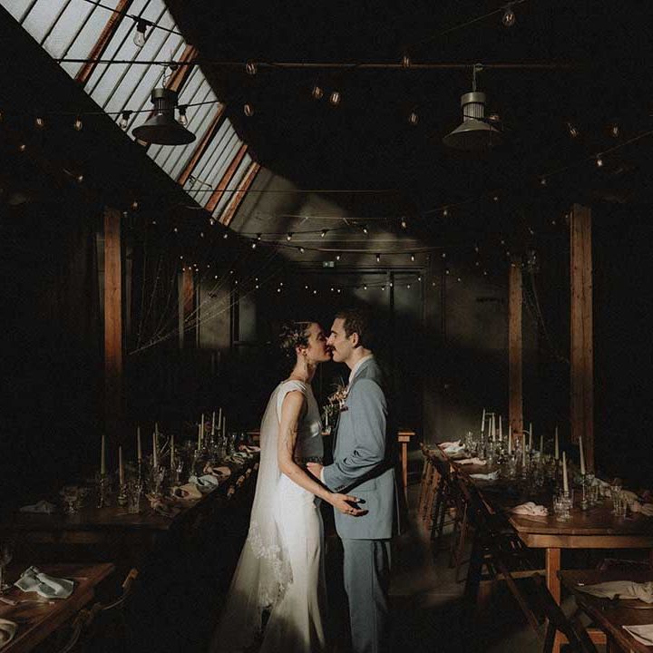 Lisa & Gabriel kiss in the middle of wooden banquet tables and industrial setting for wedding in France 